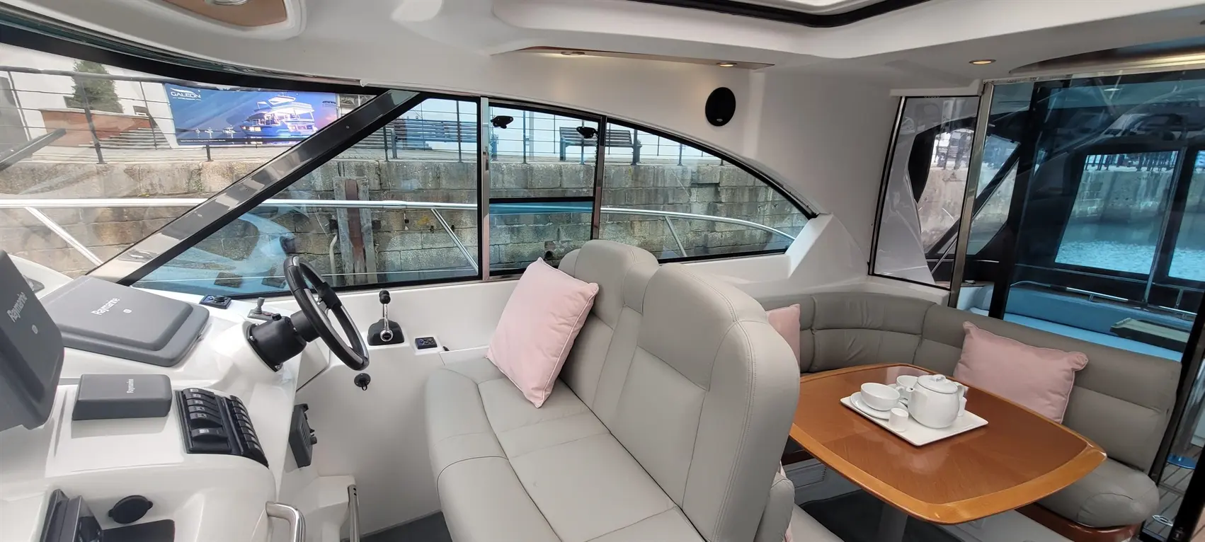 Interior of a boat featuring a steering wheel, leather seating with cushions, and a wooden table set with tea service.